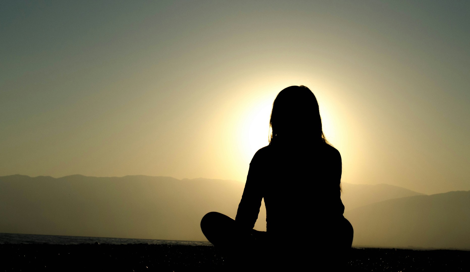 Silhouette of person meditating at sunset by mountains.