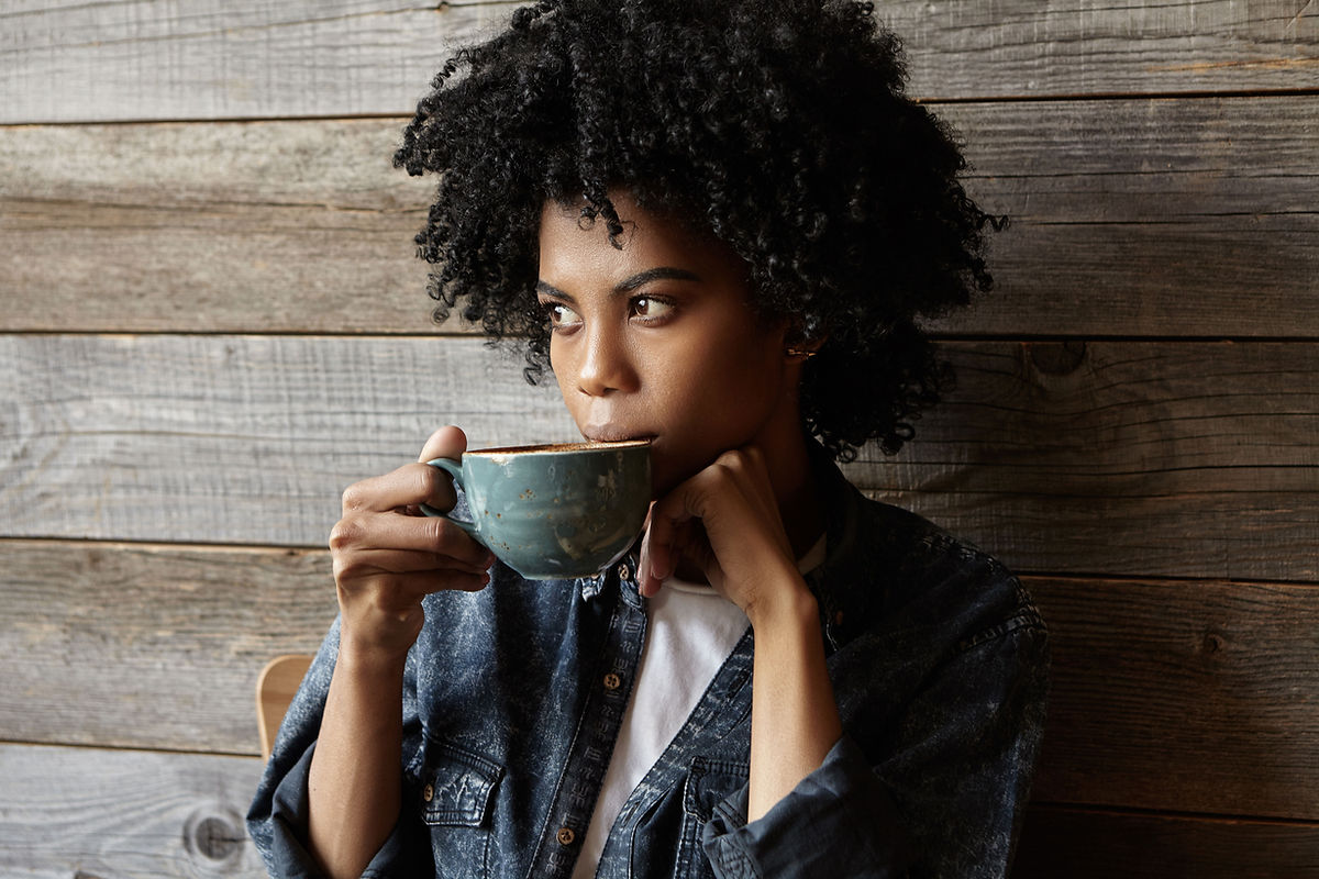 Black woman sitting alone, thoughtfully drinking a cup of coffee, appearing contemplative and reflective.
