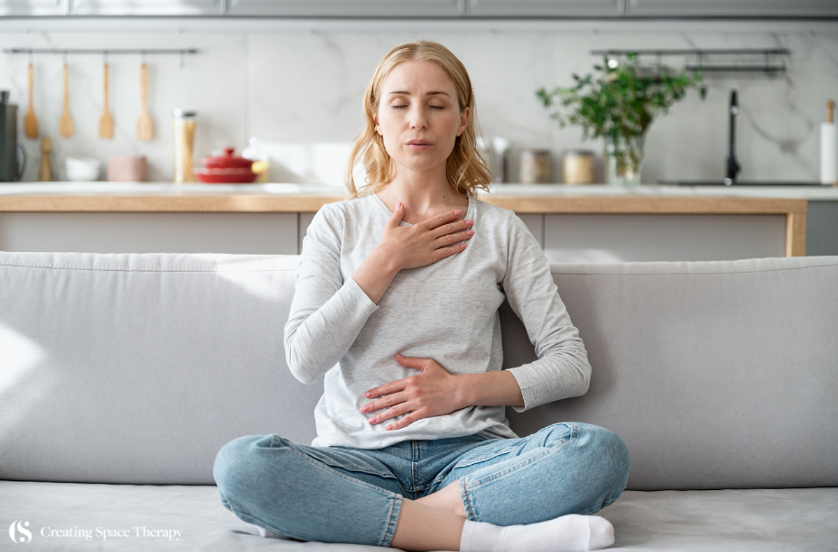 Woman meditating on a sofa with eyes closed, hands on chest and abdomen. Calm kitchen background, grey tones. Text: Creating Space Therapy.