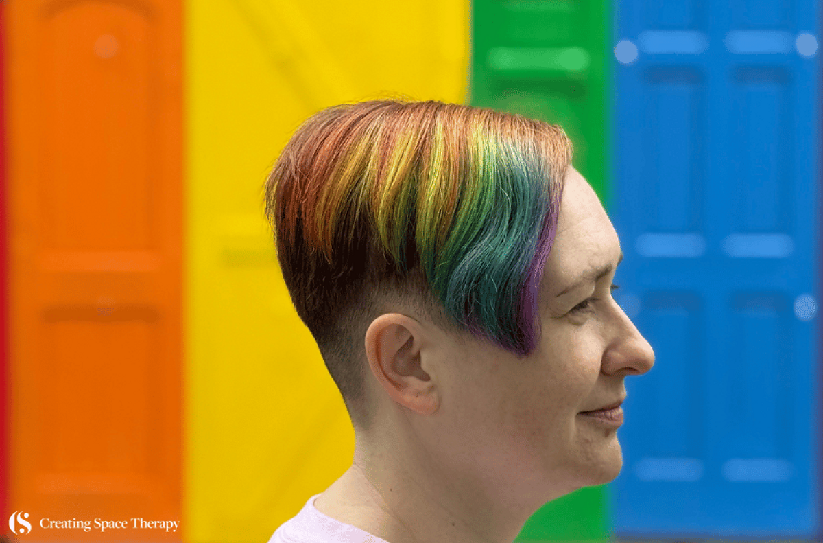 Side profile of a smiling person with rainbow-colored hair standing in front of brightly colored rainbow doors, representing LGBTQIA+ Pride and identity. Creating Space Therapy logo in the corner.