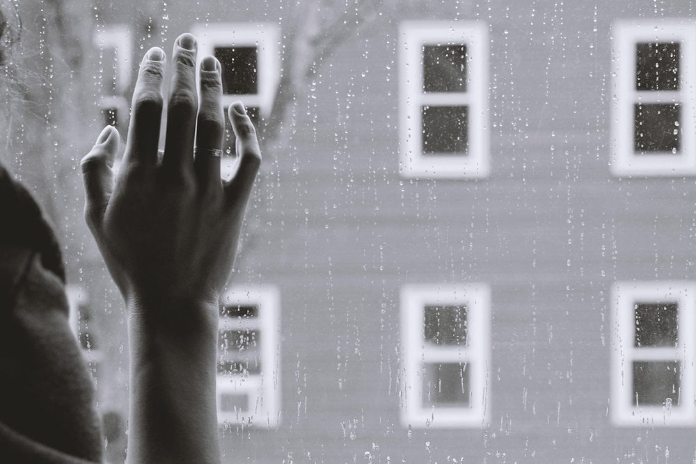 Hand reaching against rainy window, gray building outside.